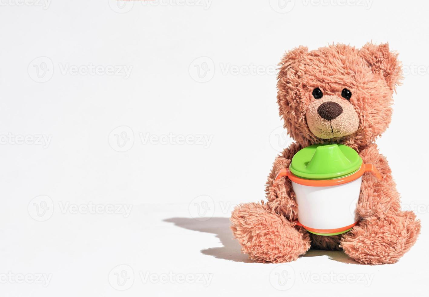 teddy bear sitting on white background and holding baby cup. kids nutrition concept, baby's first food. copy space for text. photo