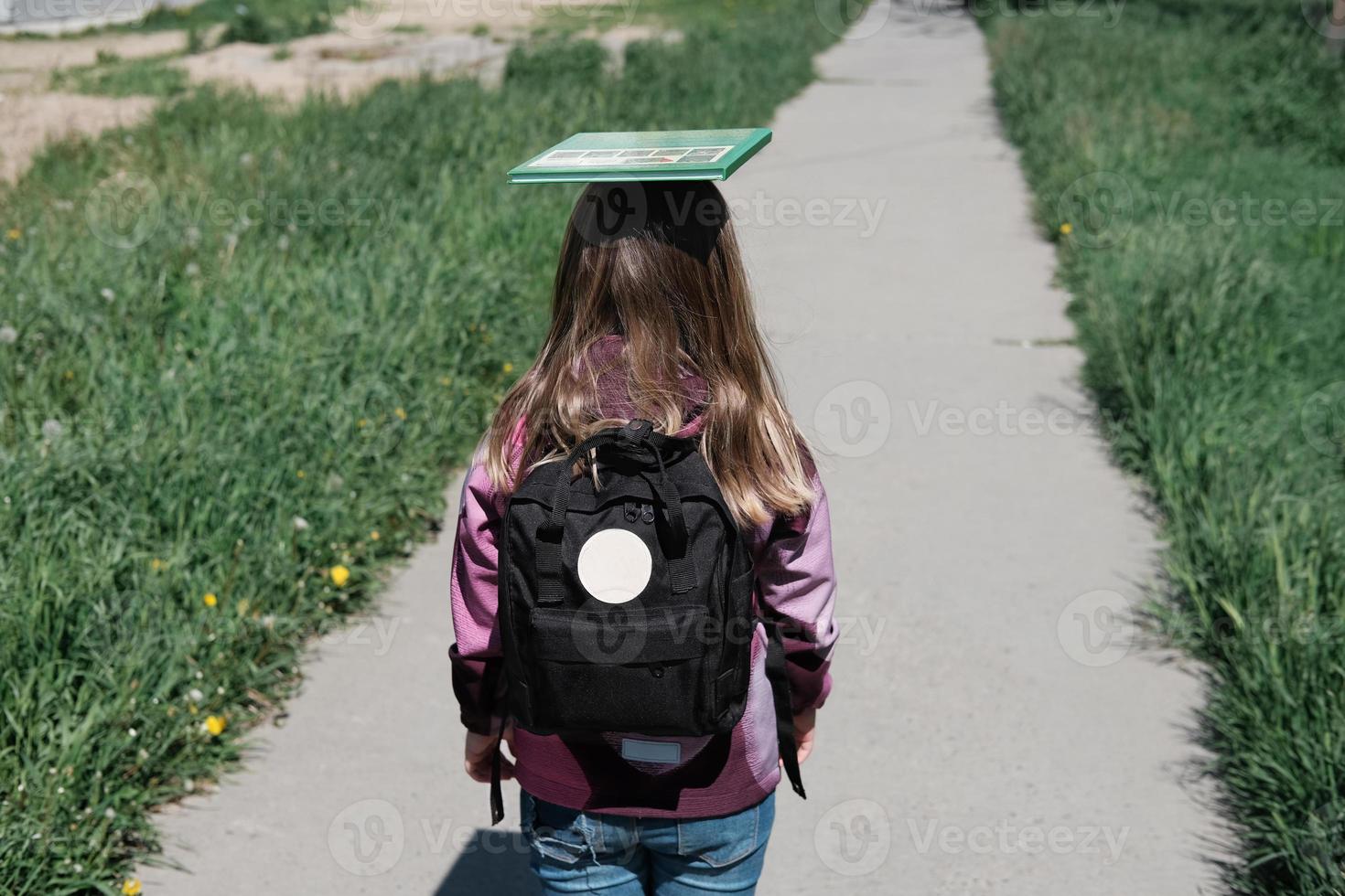 little girl going to school with schoolbag and a book on a head top. photo