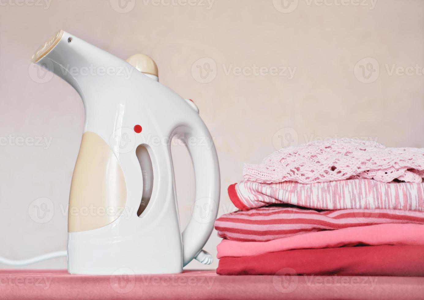 portable electric garment steamer and pile of folded kids clothes on ironing board. housekeeping and householding, chores concept. photo