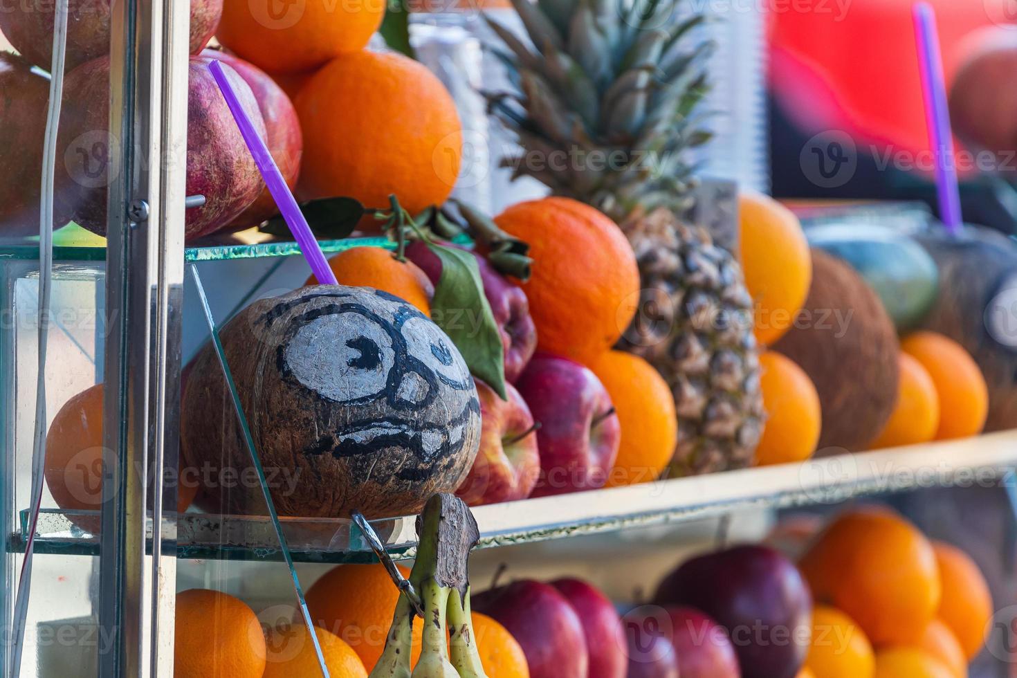 close up of a coconut with painted eyes and a smile, oranges, pomegranates and other fruits on the display of a bar with freshly squeezed juice photo