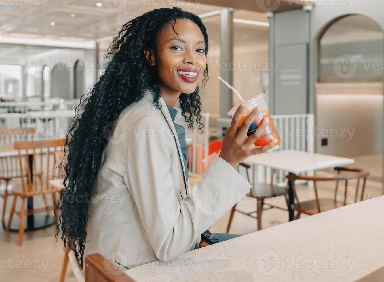 Portrait of black woman with long afro hair style at coffee bar, drinking cold beverage looking at camera smiling. Businesswoman enjoys relaxing with glass of caffeine on coffee break for refreshment. photo