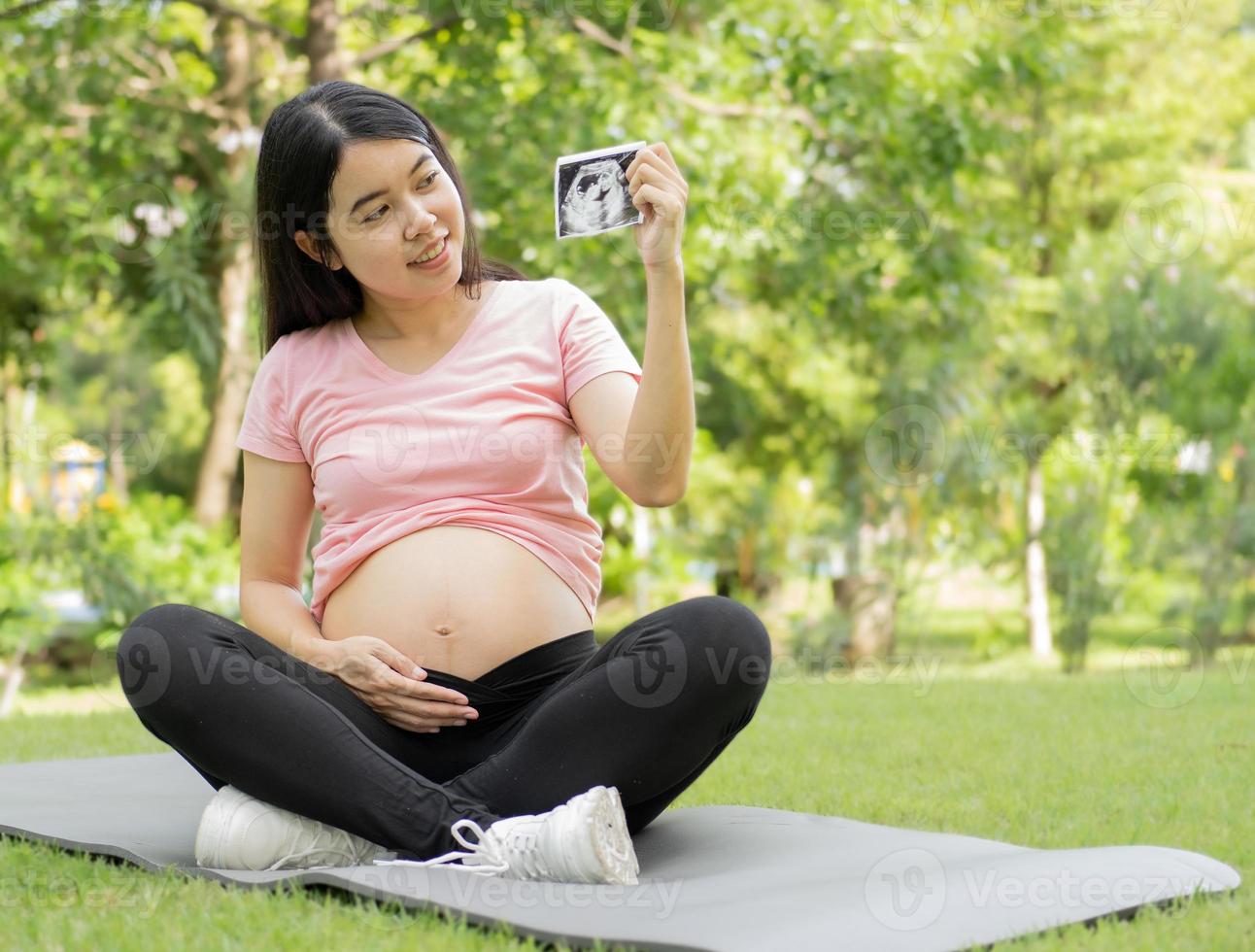 feliz mujer embarazada sentada relajándose afuera en el parque mirando una imagen de ultrasonido y sonriendo. joven asiática embarazada tocándose el vientre con cuidado y amor. concepto de embarazo y maternidad. foto