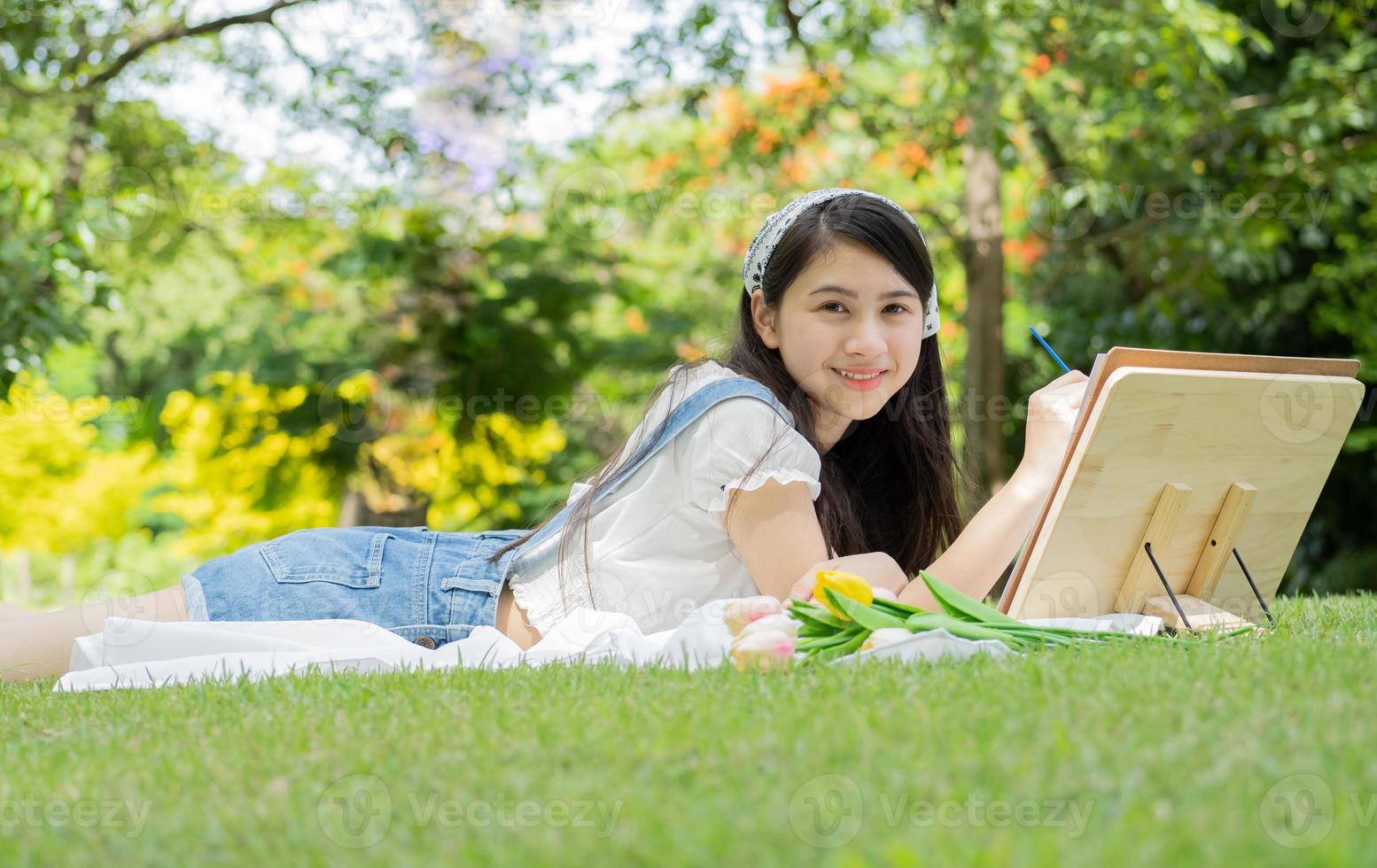 Portrait of young Asian woman lying on green grass in city park outdoor painting with a brush. Blurred bushes and trees are the nature background. Smiling teenager artist, painter relaxing as drawing. photo