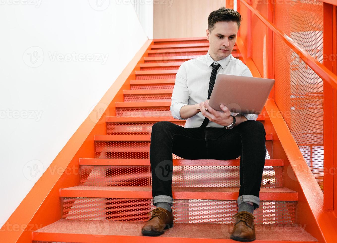 Young businessman sitting on red stair using laptop computer in modern office. Concentrated man using notebook. Handsome white collar worker looking at laptop screen during internet remote meeting. photo