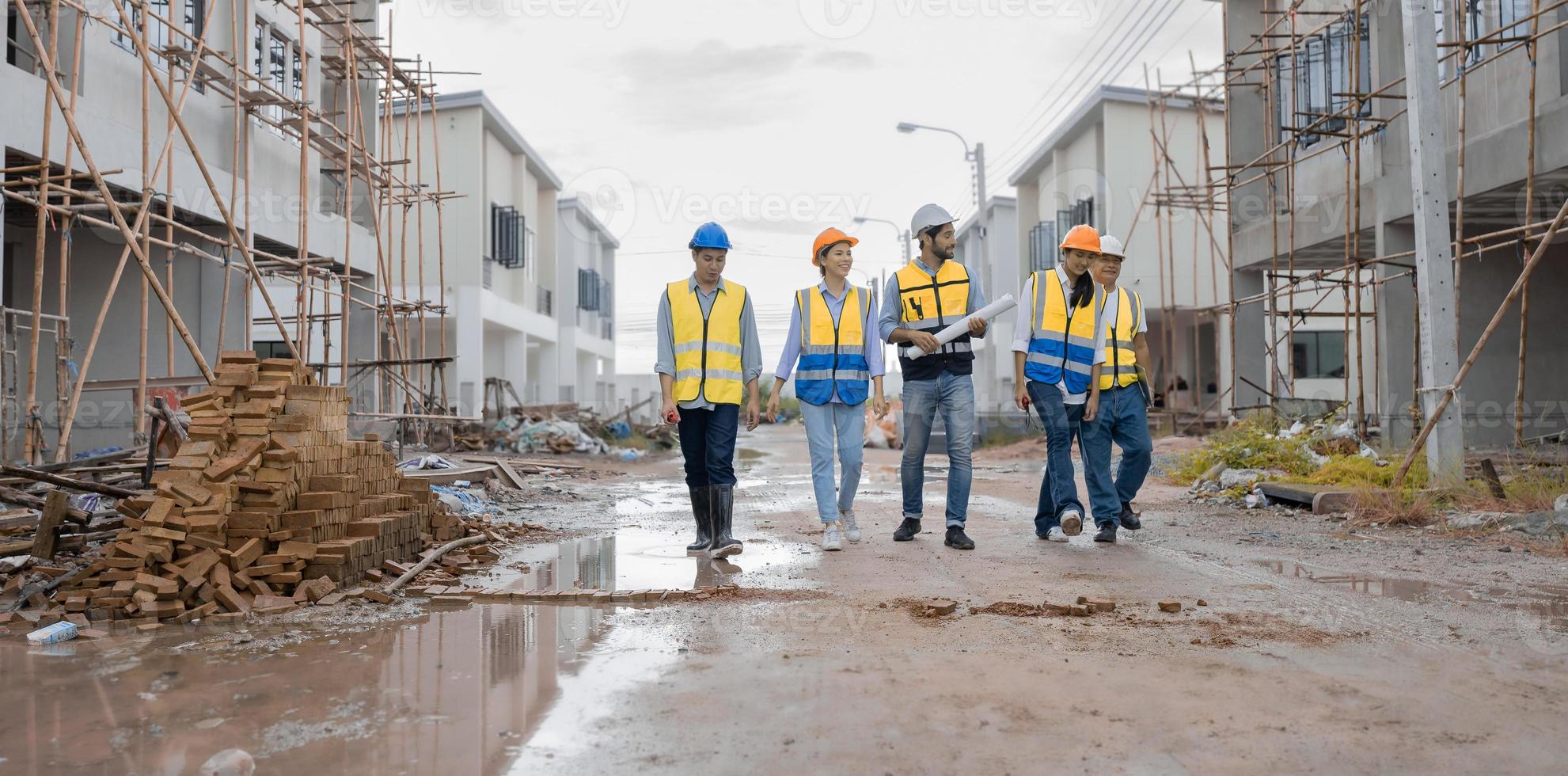 Team of engineers and workers wear safety clothing are walking through wet street in residential housing construction site. Engineering teamwork talking, discussing building project development. photo