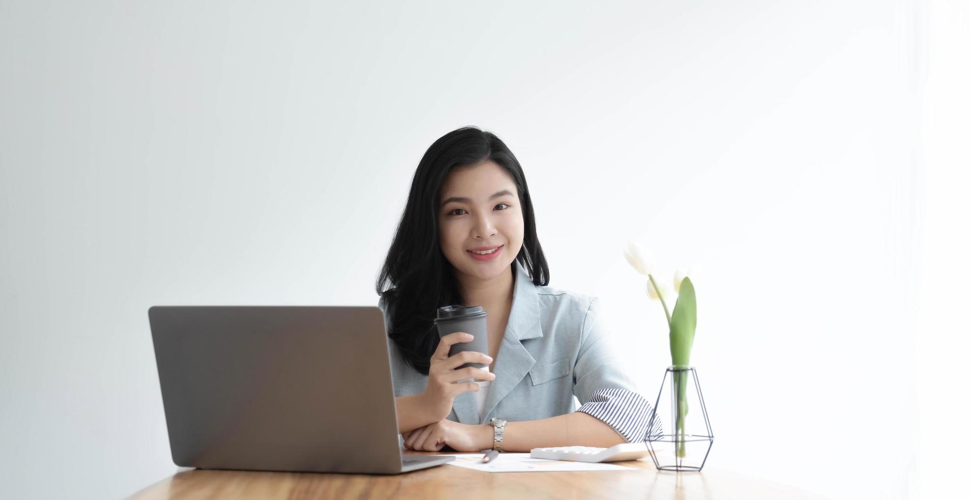 Smiling Asian businesswoman holding a coffee mug and laptop at the office. Looking at the camera. photo