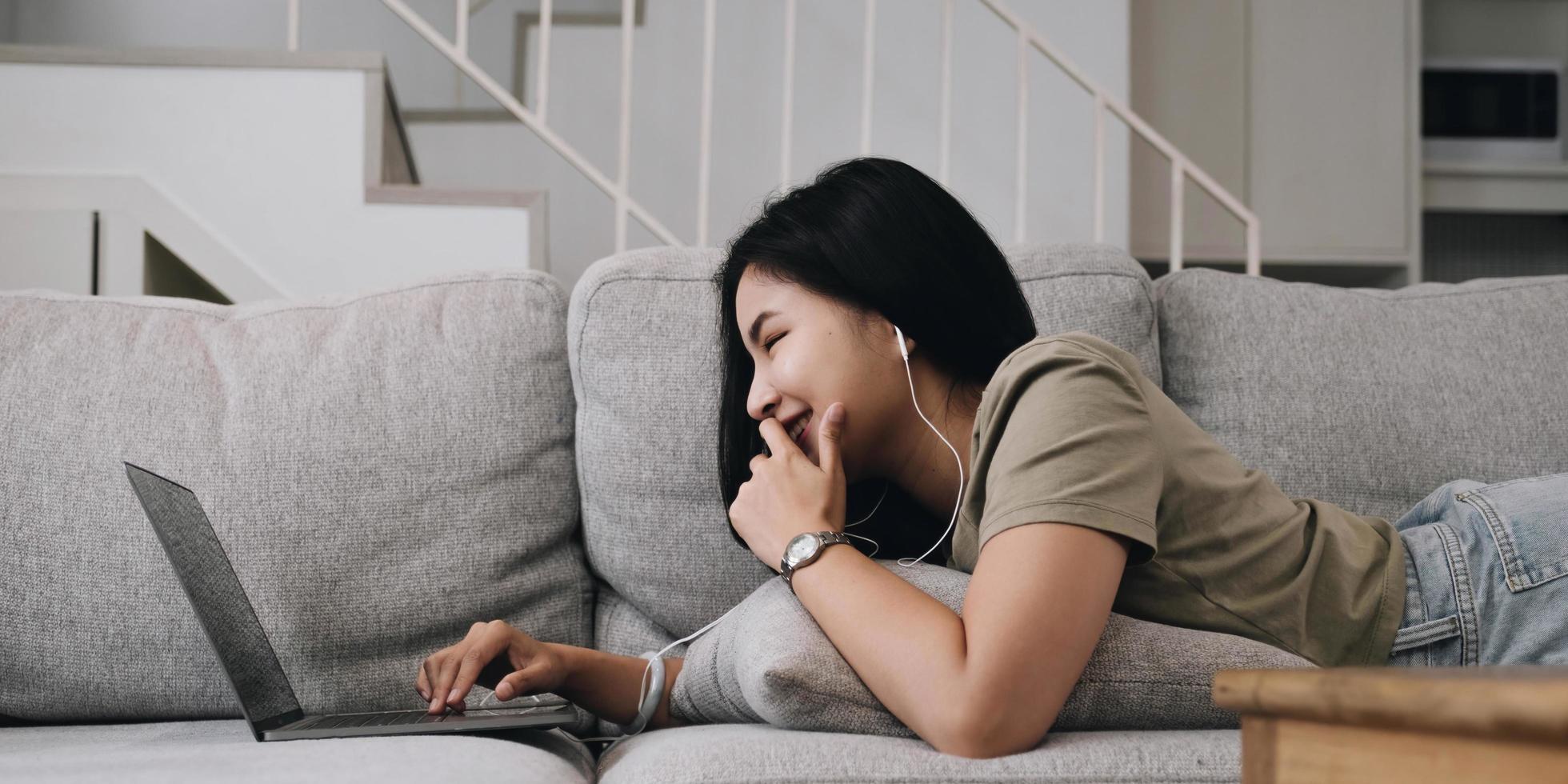 Asian woman lying on the sofa in the living room listening to music on a laptop device woman relaxing on the sofa at home View messages on the computer screen on the device. communication concept photo