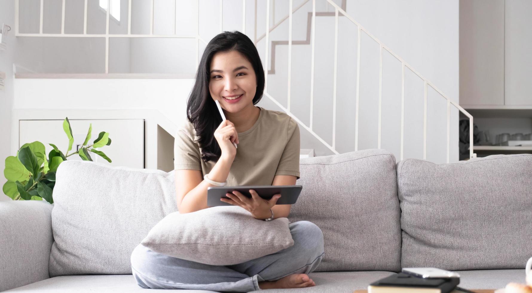 Beautiful smiling Asian woman looking at camera relaxing on leather sofa at home. work on a laptop computer photo