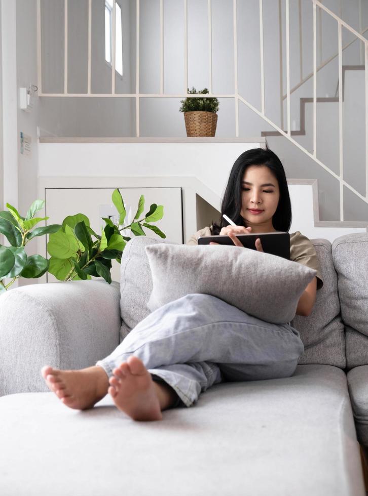 Attractive smiling young asian  woman relaxing on a leather couch at home, working on laptop computer. photo