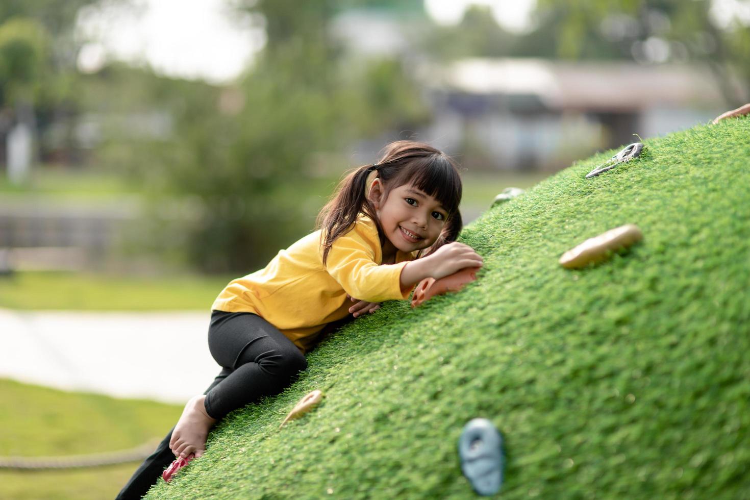 linda chica asiática divirtiéndose tratando de escalar rocas artificiales en el patio de la escuela, niñita trepando por la pared de roca, coordinación mano-ojo, desarrollo de habilidades foto