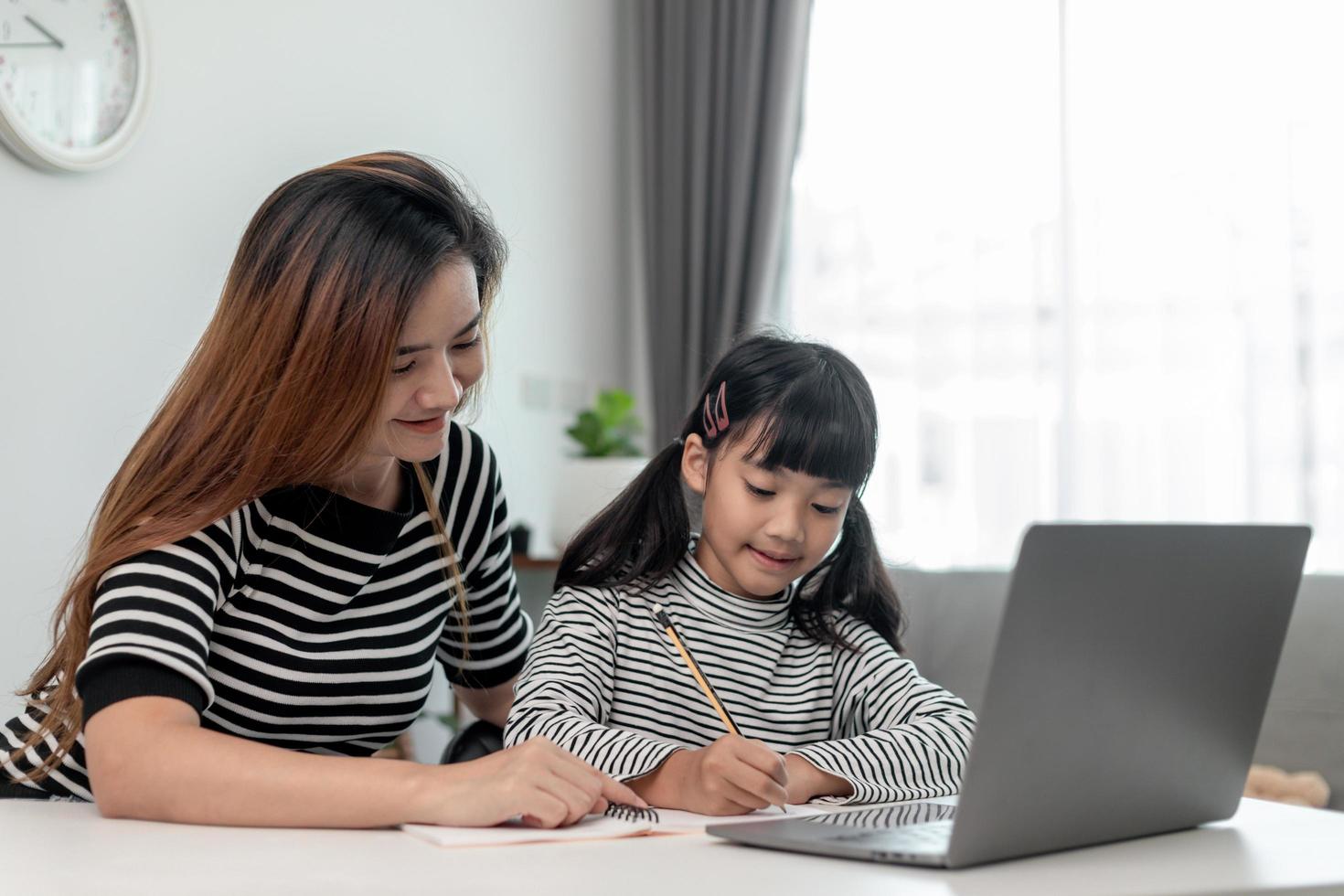 Asian little young girl kid learning online class at home with mother. Preschool child use laptop computer do homework, homeschool from school teacher by digital remote internet with support from mom. photo