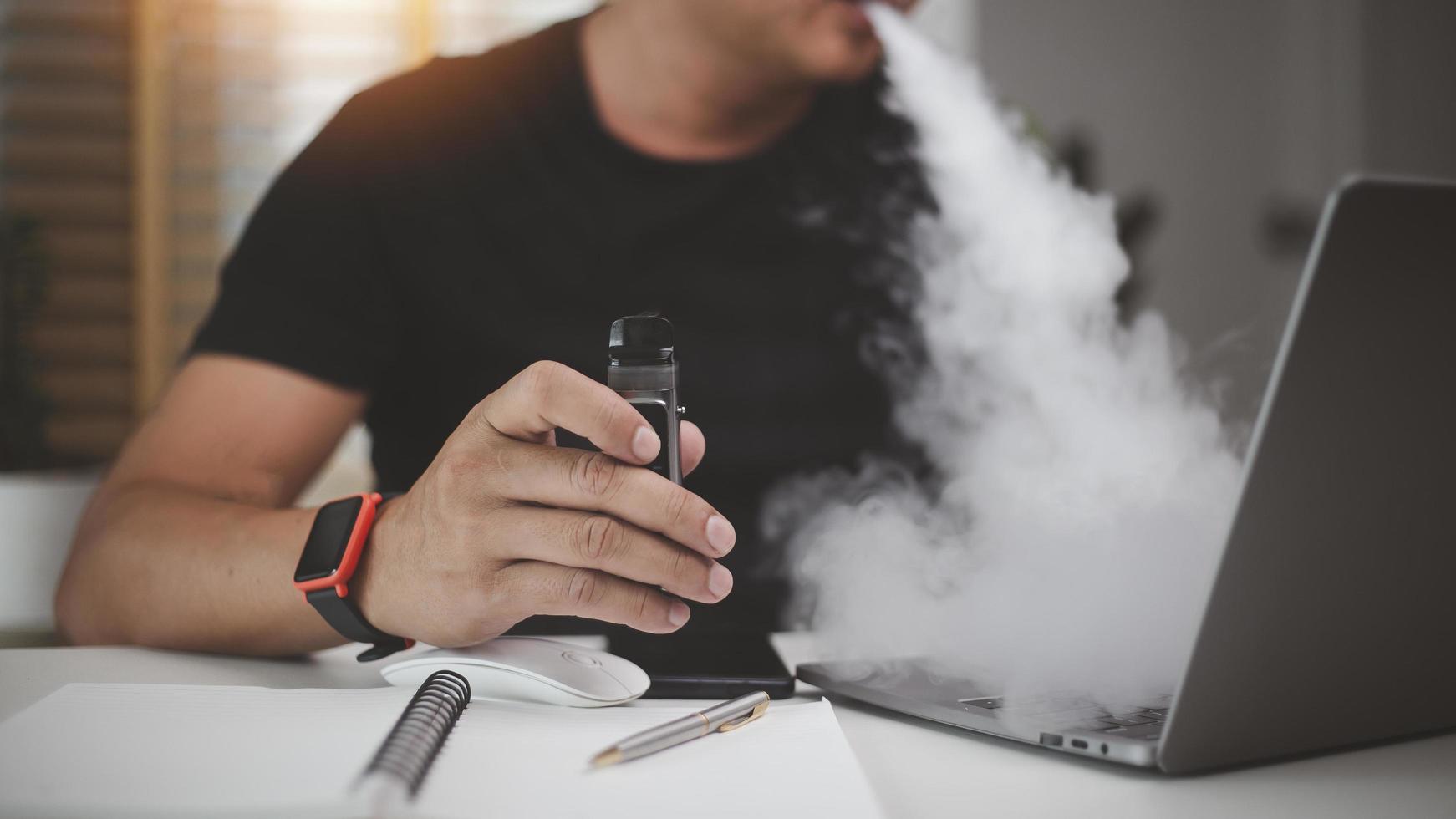 young man using disposable electronic cigarette at table in office photo