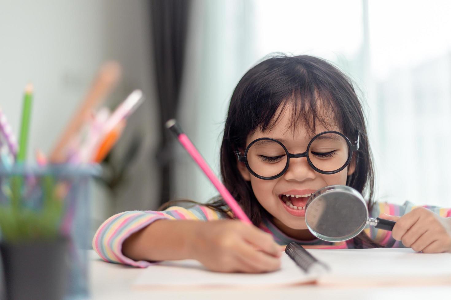 Student little Asian girl writing with a magnifying glass looking at the camera. sweet kids sitting in the living room at home. family activity concept. photo