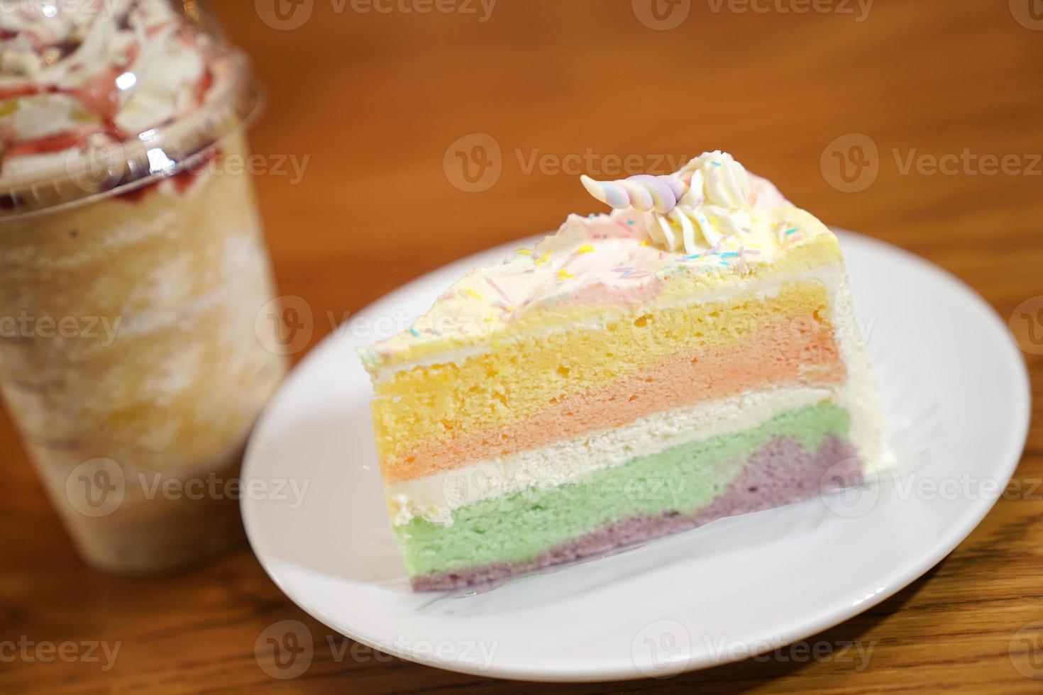 Slided rainbow cake decorate with unicorn horn on top next to coffee frappe. The cake is in a white disk on the wooden table and blur background. photo