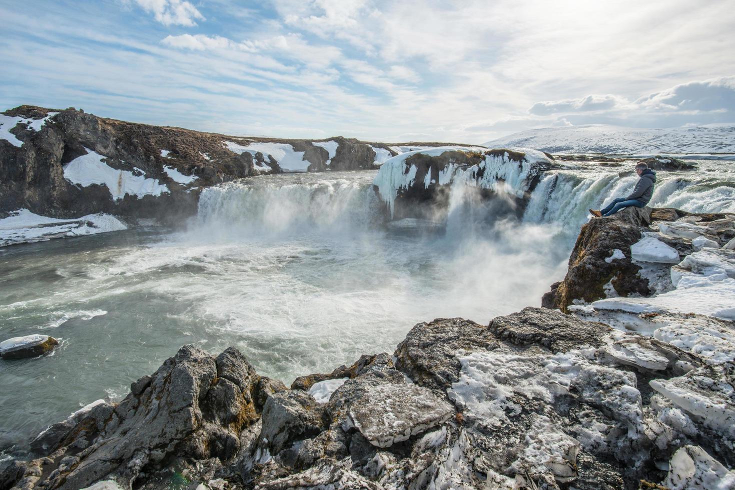 turista europeo sentado en el borde del acantilado frente a godafoss, la cascada de dios, uno de los lugares de atracción turística en la región norte de islandia. foto