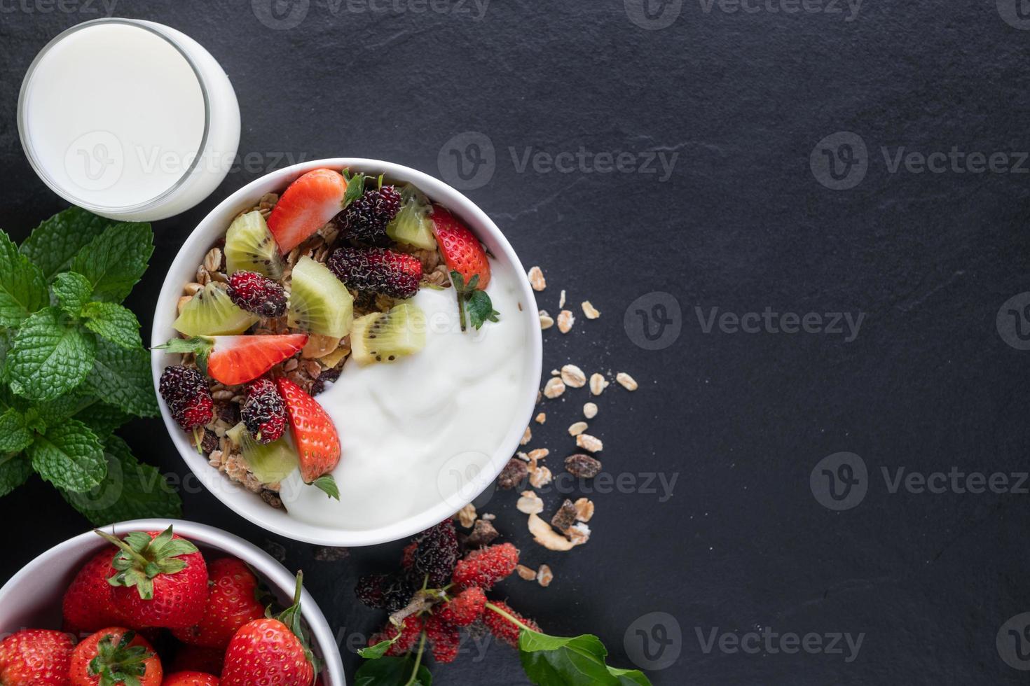 bowl of oat granola with yogurt, fresh mulberry, strawberries, kiwi mint and nuts on the black rock board for healthy breakfast, top view, copy space, flat lay. Healthy breakfast menu concept. photo