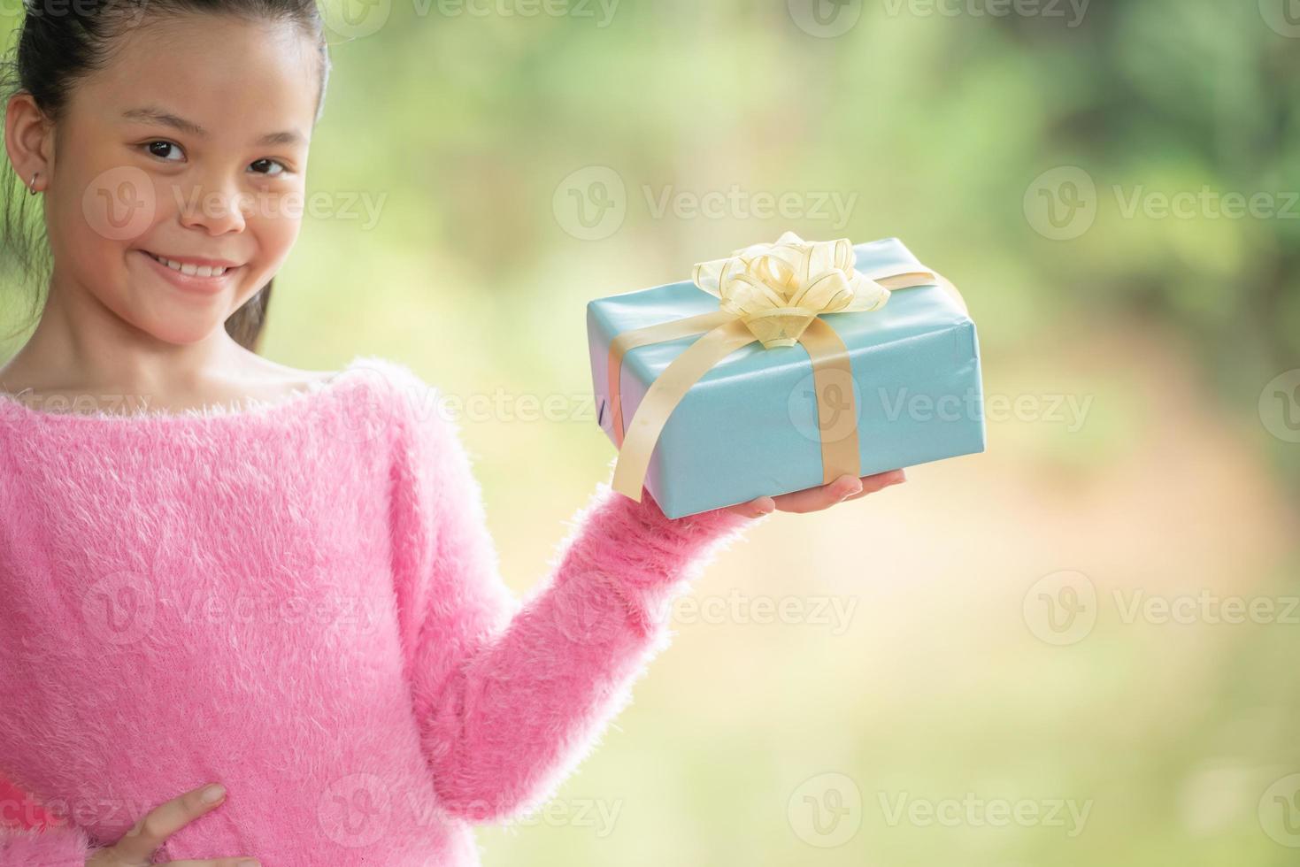 Christmas portrait of happy smiling little girl child with gift box near a green branch tree. green leaves bokeh out of focus background from nature forest. photo