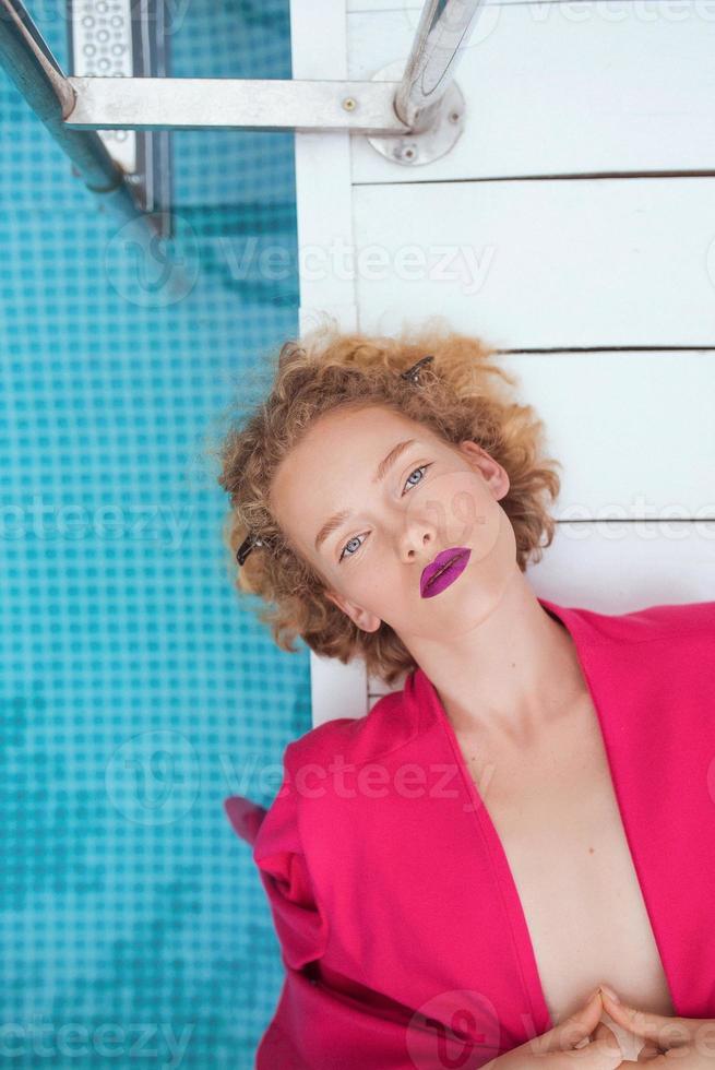 portrait of marvelous stylish redhead curly young woman laying in fuchsia jacket by the swimming pool. Fashion, style, leisure, recreation, travel, make up concept. Film grain photo