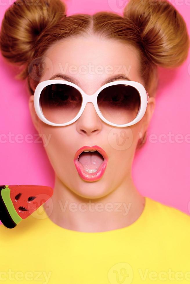 Portrait of stylish cute young blonde woman in sunglasses with colorful watermelon lollipop on pink background photo