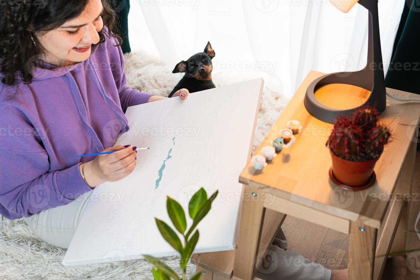 The adult woman painting by numbers indoors, her pet miniature pincher near. People with pets. photo