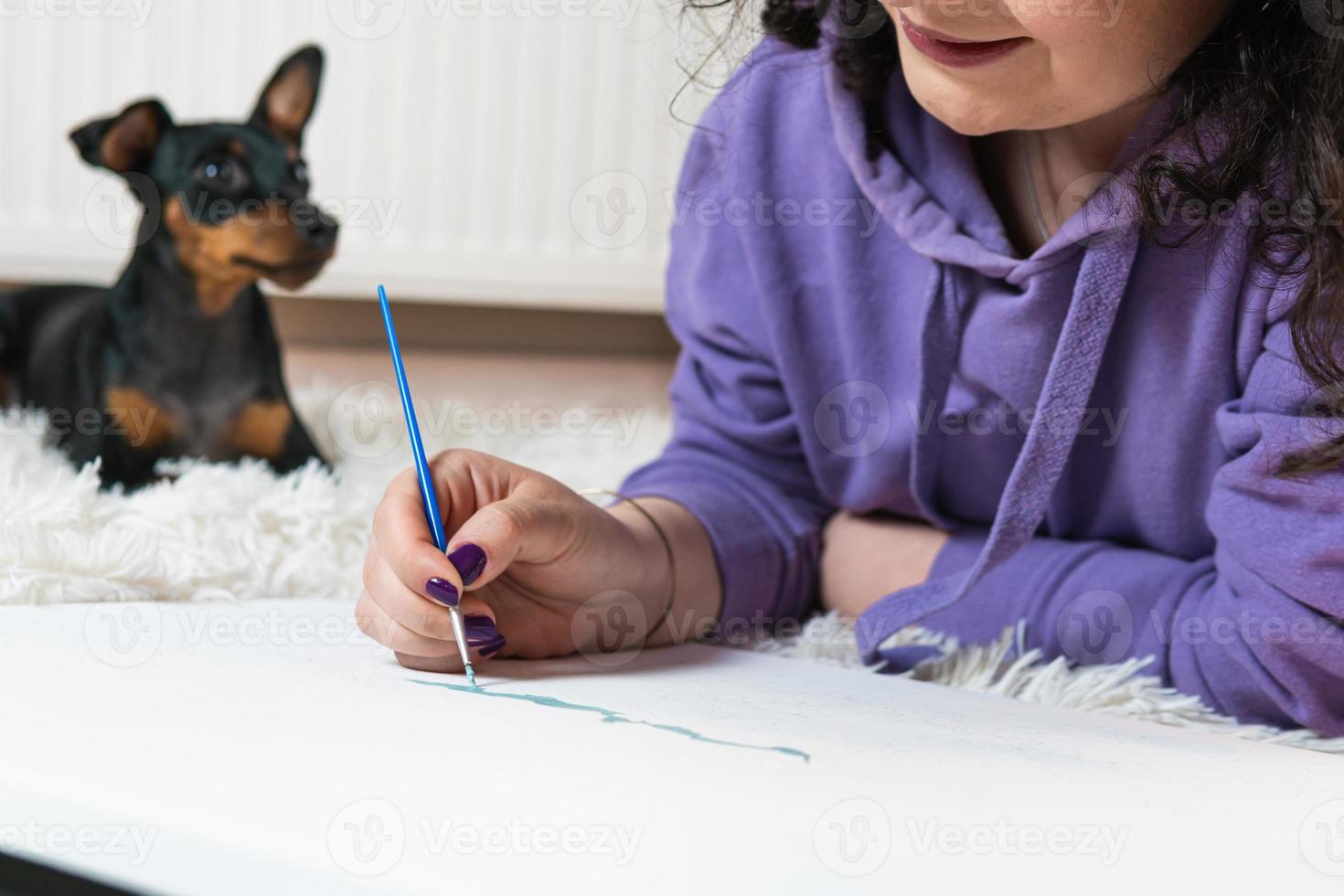 The adult woman painting by numbers indoors, her pet miniature pincher near. People with pets. photo
