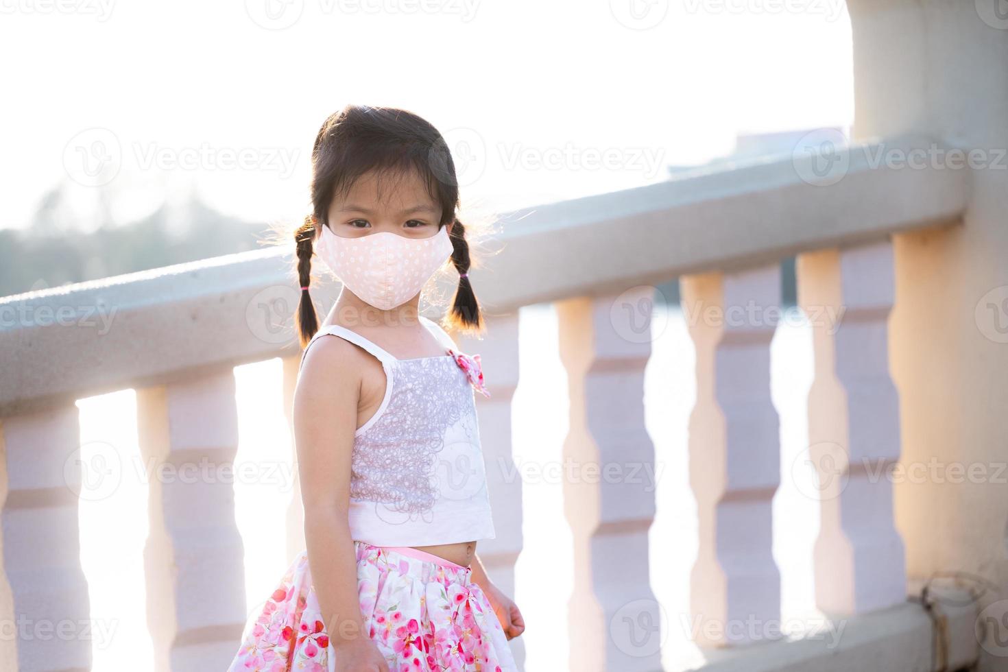 Portrait adorable kid girl wearing cloth face mask travel public park in evening. Summer or spring time. Child stand and watch nature at foot of bridge across pond. Beautiful children aged 5 years old photo