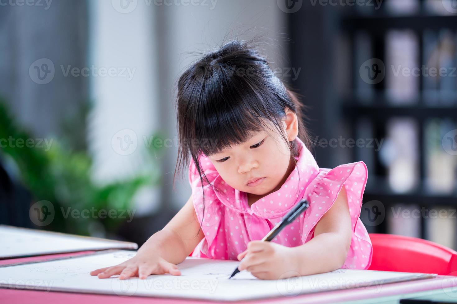 Portrait Asian kid sitting on large sheet of drawing paper on table. Students concentrate on art work. 4 year old girl wears pink shirt with white polka dots. Kindergarten child in homeschool class. photo