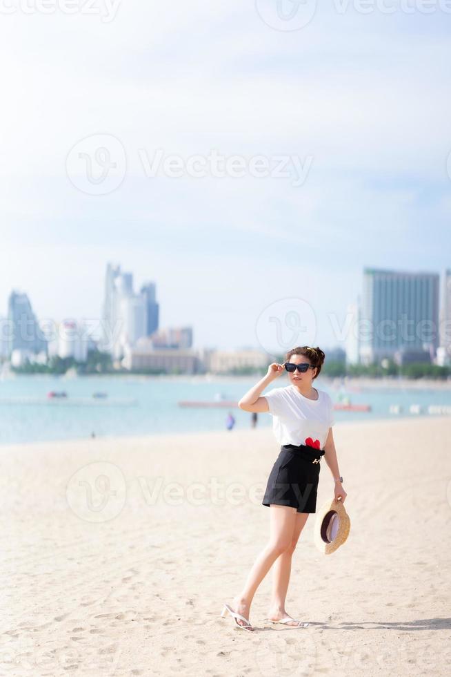 adolescente de pie en la playa mirando hermosos paisajes, sosteniendo sombrero de paja y gafas de sol, sonriendo alegremente. durante el verano o la primavera. en un cómodo día de viaje. espacio vacio. imagen vertical foto