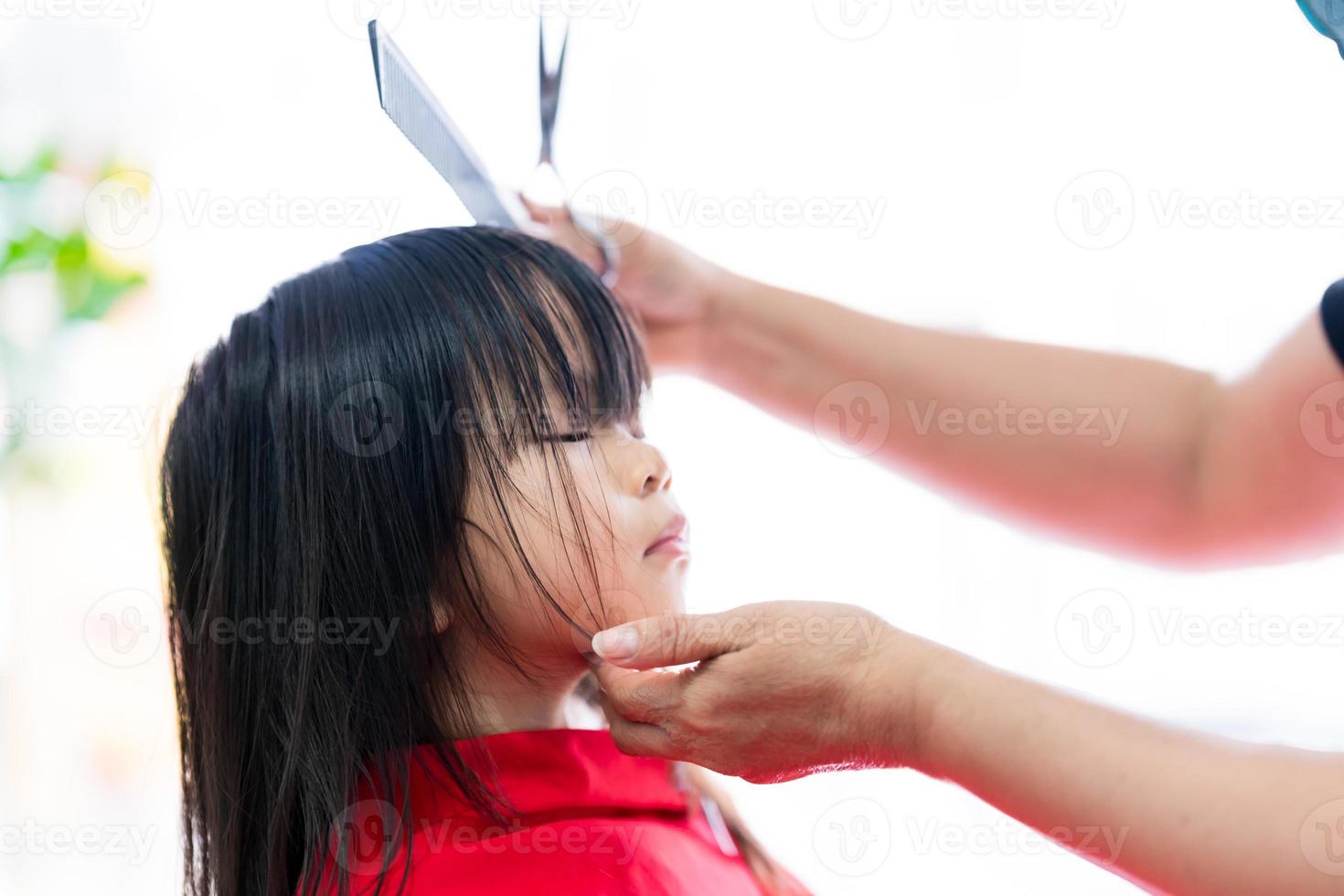 Portrait of child 3-4 years old. Cute Asian girl sits still for barber to style hair beautifully. Beauticians hands hold comb and scissors and gently support her little face. photo