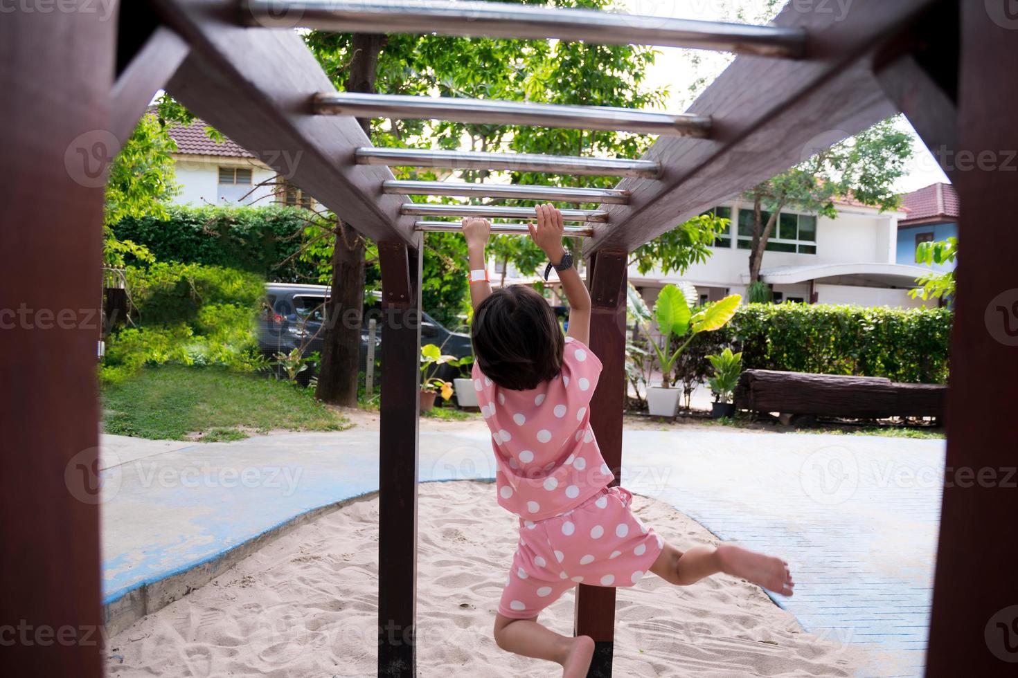 Rear back view. Preschool kid girl is hanging out on bar at playground. Healthy children exercise by having fun. Empty space for entering text. Portrait image 6 year old child wears pink shirt. photo