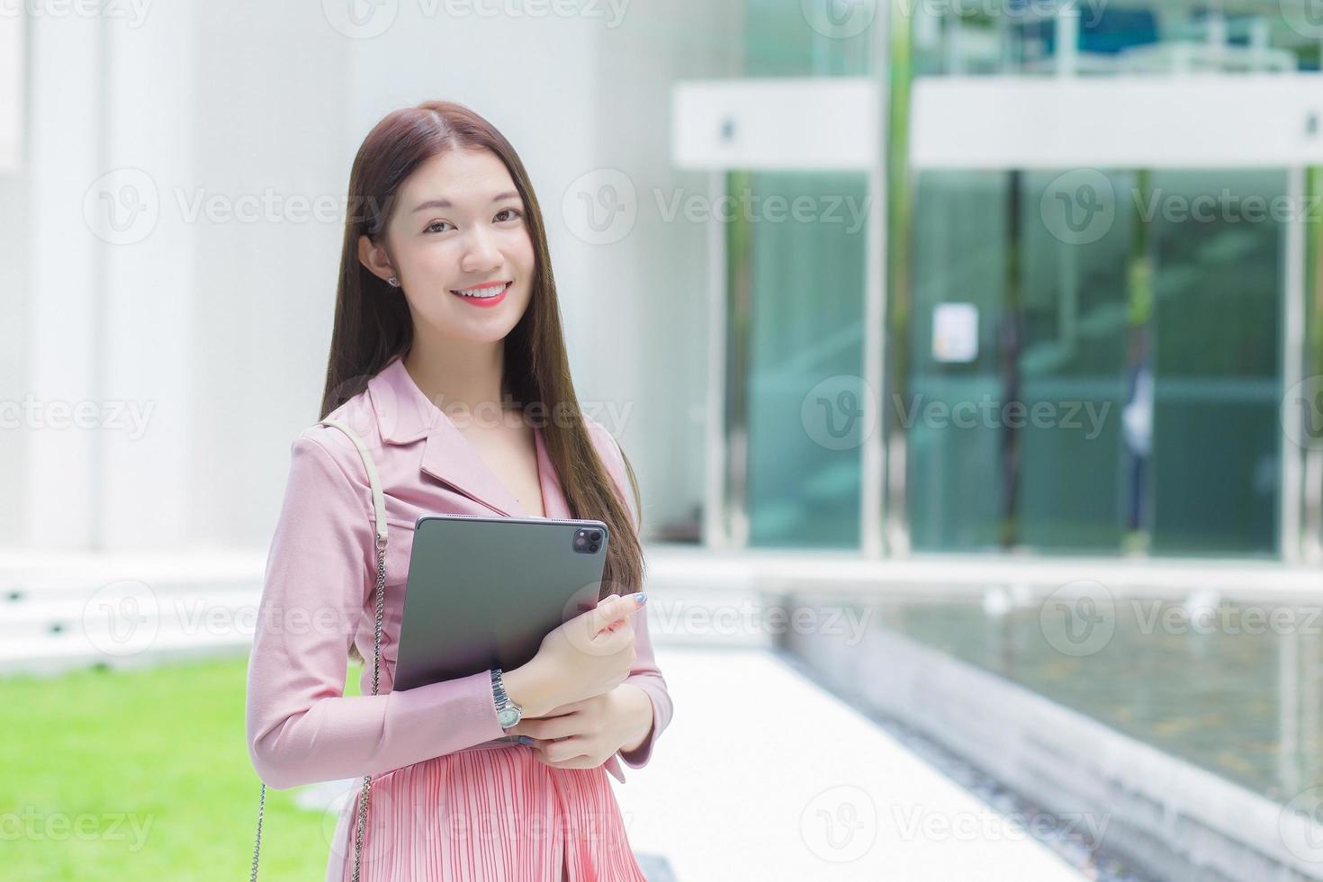 Asian professional business female with long hair is smiling outdoor in the garden front of the office while hold at the tablet in her hand to go out meeting with clients at the office. photo
