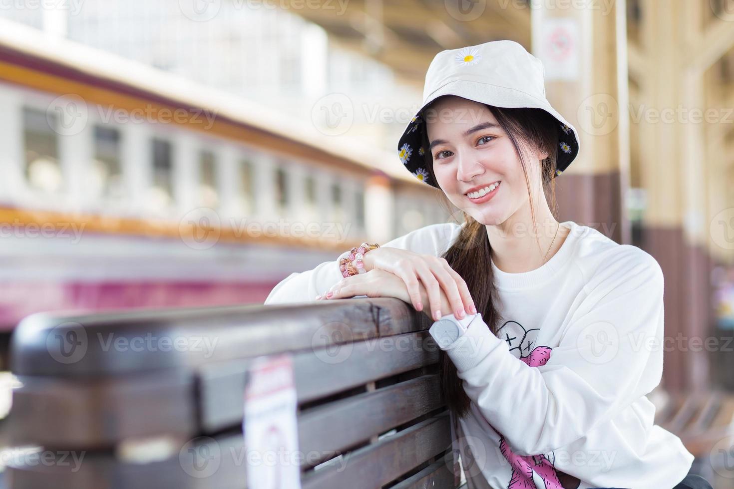 Asian beautiful woman in a long-sleeved white shirt and a hat sits happy smilie in the train station waiting for the train to arrive. photo