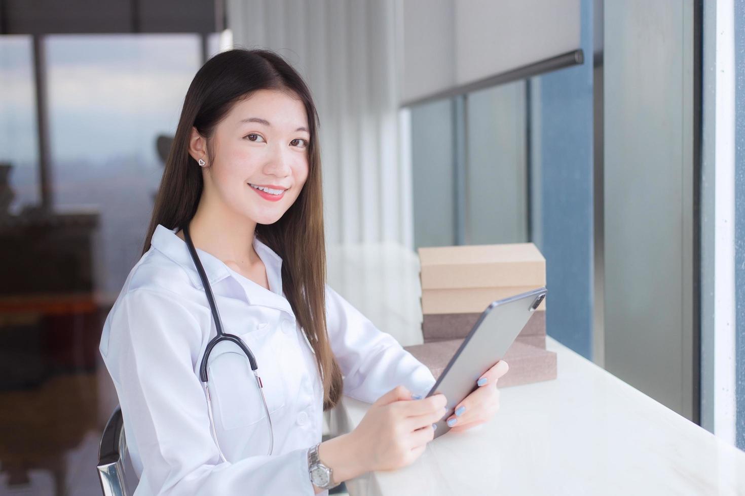 Professional Asian female doctor dressed in white medical coat sits on chair at a hospital library to search some information on a tablet for treatment. photo