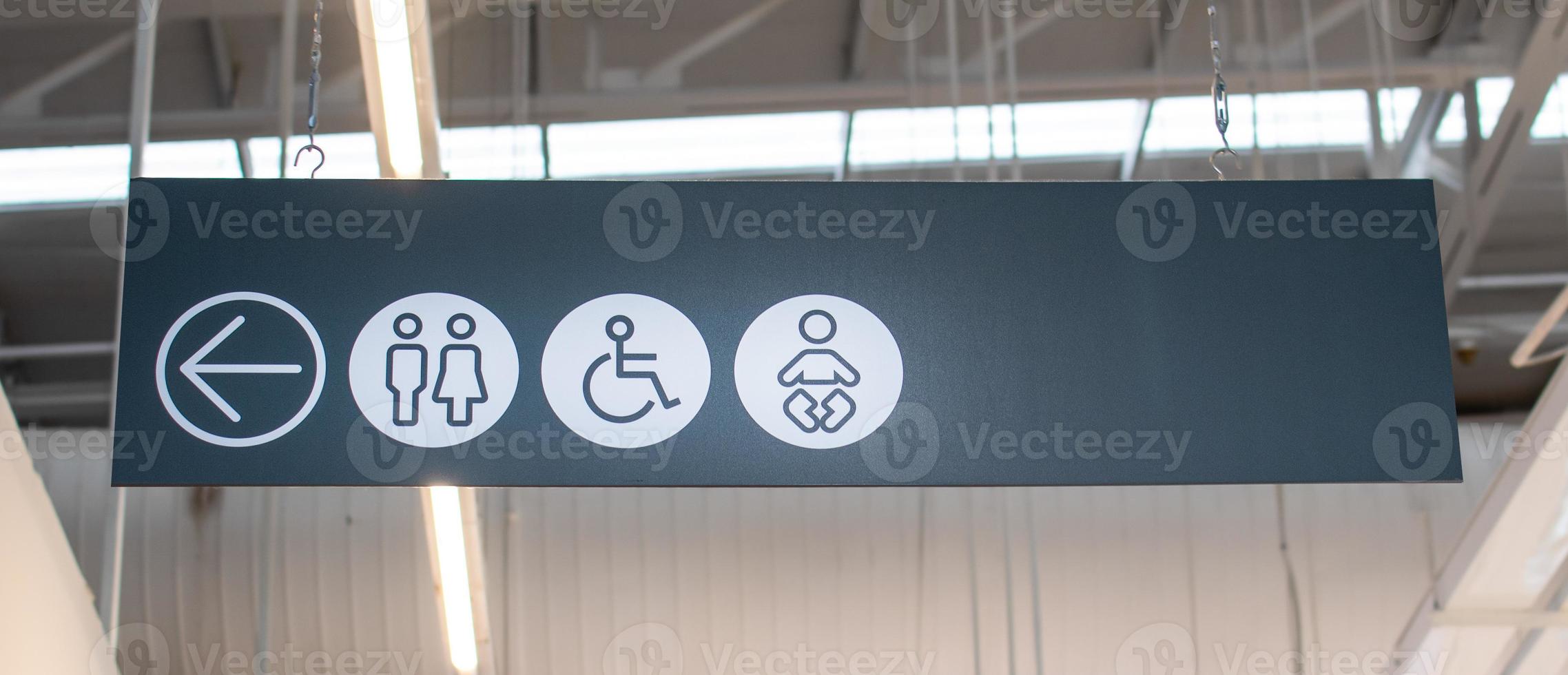 Sign to the bathroom on the left is a black and white symbol is hung to see. The sign contains arrow symbols to the left, women, men, disabled people, and children. Selective focus. Blurred background photo