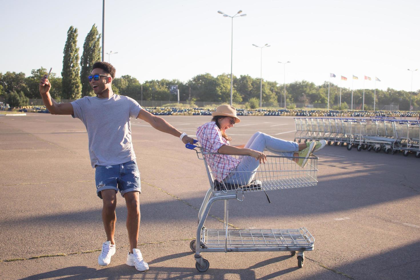 Happy young couple riding on trolley on empty mall parking , hipster friend have good time during shopping, couple in love riding on shopping cart photo
