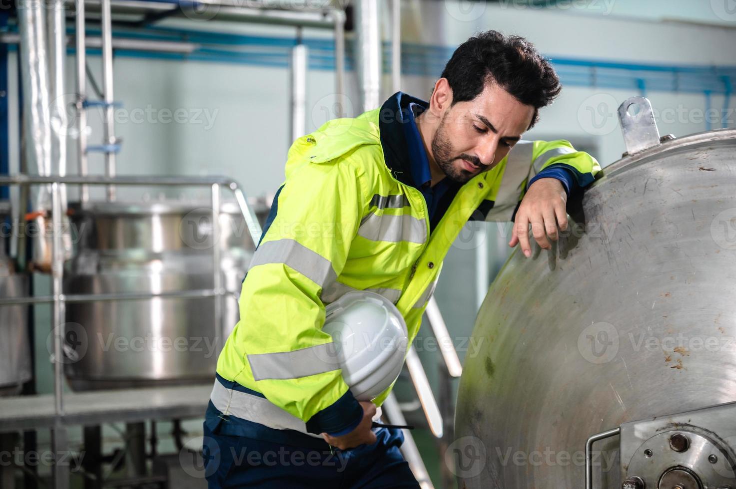 Young male machine inspector wearing vest and hardhat with headphones checking machine and sterilizers in water plant while making notes in digital tablet photo
