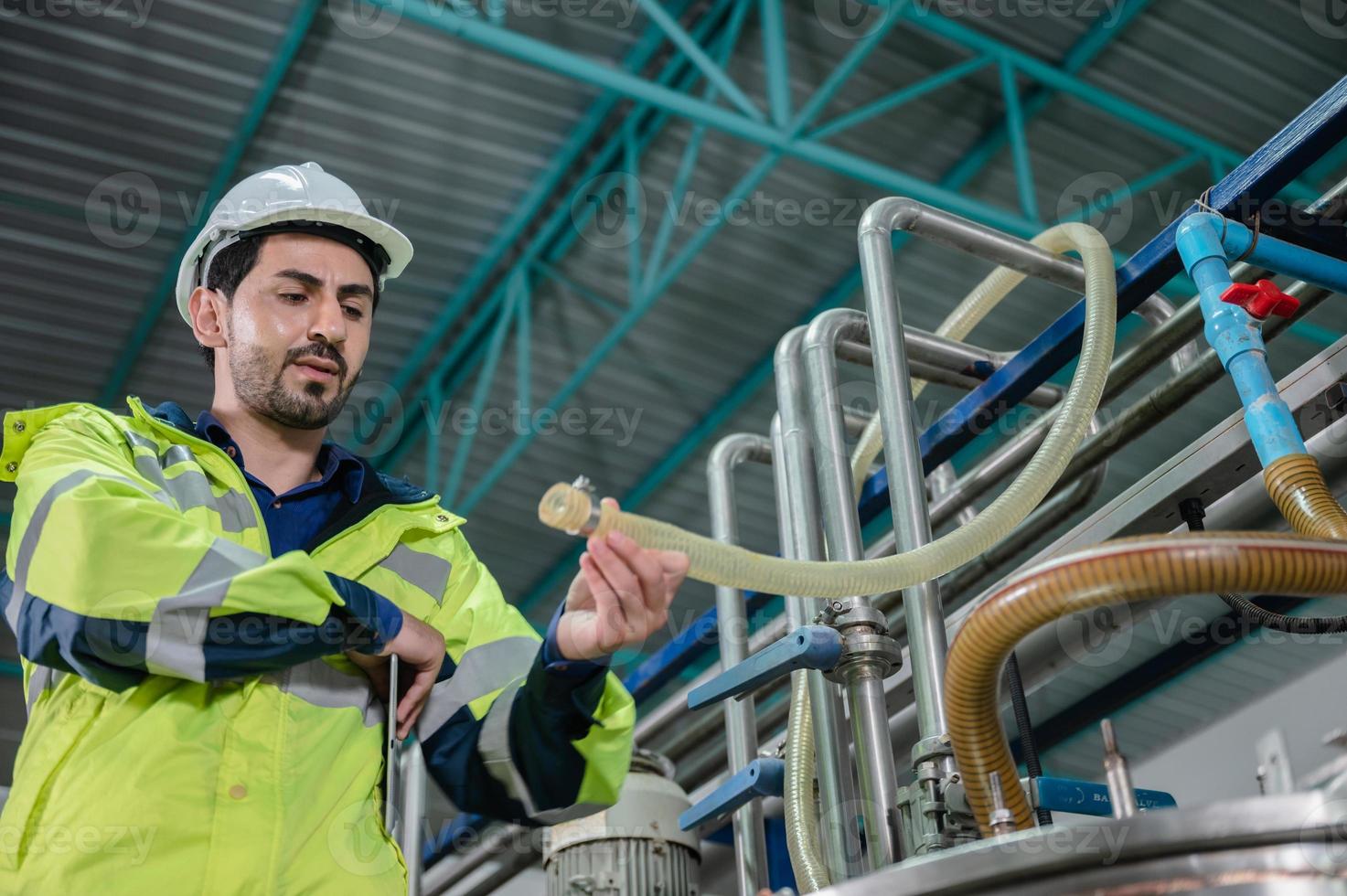 Young male machine inspector wearing vest and hardhat with headphones checking machine and sterilizers in water plant while making notes in digital tablet photo
