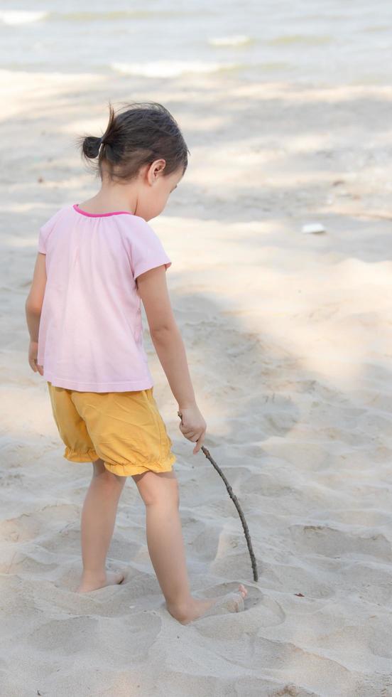 Positive charming 4 years old cute baby Asian girl, little preschooler child writing and drawing on the sand beach with little wood stick on beautiful sunny day in summer time. photo