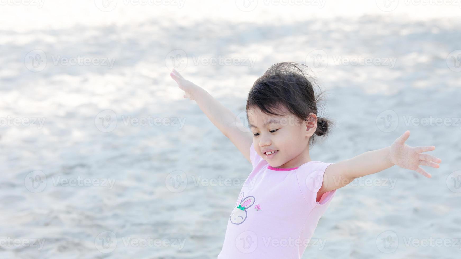 Portrait of happy charming 4 years old cute baby Asian girl, little preschooler child smiling and spreading hands, have fun on white sand background. Freedom concept. photo