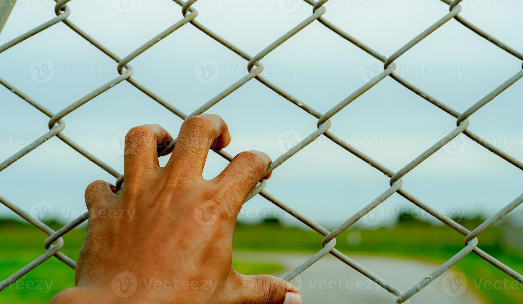 Man hand holding metal chain link fence. Refugee and immigrant concept. Life and freedom. Anguish, gloom, and persecution feeling. Man hand grabbing cage or camp fence. Prison fence. Barrier border. photo