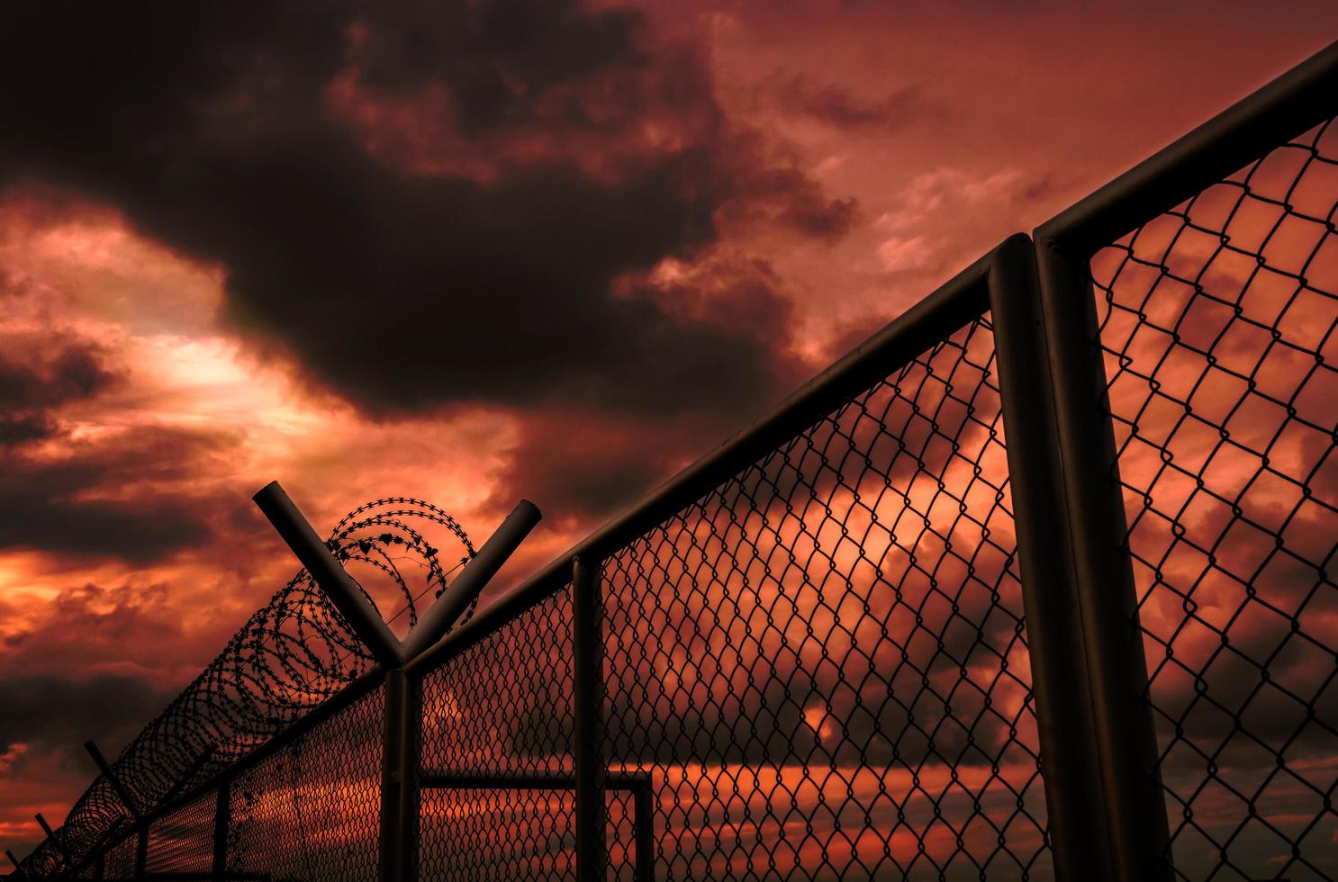 Security fence of military zone or private area fence with red sky and dark clouds. Barbed wire security fence with stormy sky. Barrier border. Boundary security wall. Dramatic and horror sky. photo