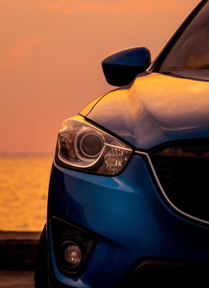 vista frontal de un camión suv azul con diseño deportivo y moderno estacionado en una carretera de hormigón junto al mar al atardecer por la noche. primer plano de la luz del faro del coche azul. concepto de tecnología de automóviles híbridos y eléctricos. foto