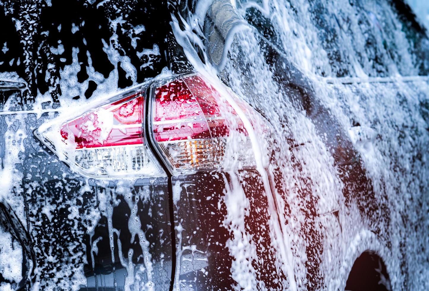 Rear view of black compact SUV car with sport and modern design are washing with water and white foam. Car care service business. Car covered with white foam. Car wash before and after travel concept. photo