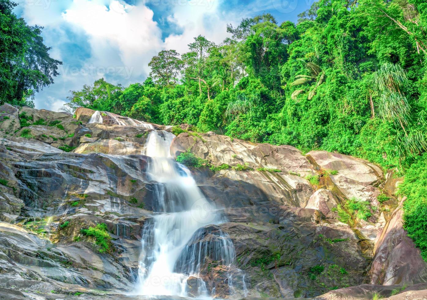 Beautiful waterfall at the mountain with blue sky and white cumulus clouds. Waterfall in tropical green tree forest. Waterfall is flowing in jungle. Nature abstract background. Granite rock mountain. photo