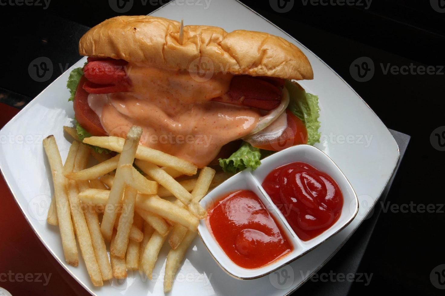 Complete hotdog package. Red sausage, cheese sauce, tomato wedges, lettuce. Plus french fries and a container of red chili and tomato sauce. Served on a white square plate. Close up. top view photo