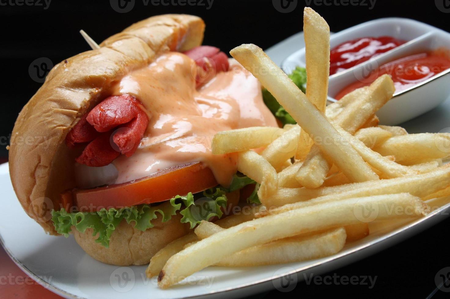 Complete hotdog package. Red sausage, cheese sauce, tomato wedges, lettuce. Plus french fries and a container of red chili and tomato sauce. Served on a white square plate. Close up. top view photo