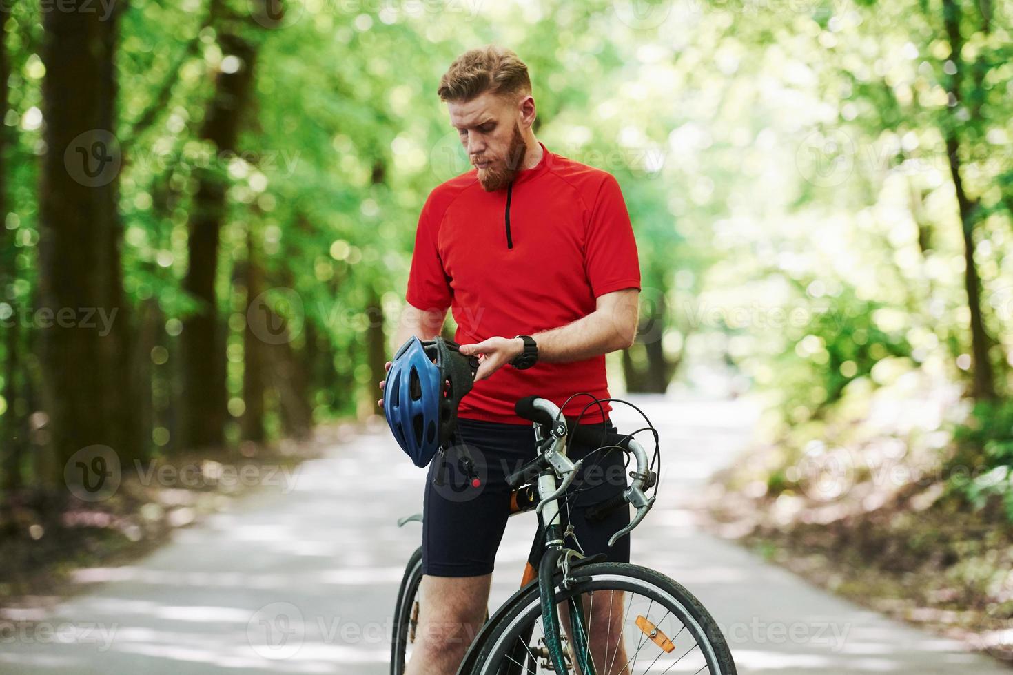 Taking off protective helmet. Cyclist on a bike is on the asphalt road in the forest at sunny day photo