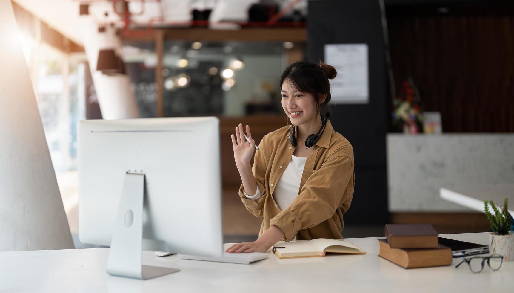 Joyful young asian female in wireless headphones waving wand greeting to screen while sitting at table and having video chat with business partners using laptop against of comfortable office photo