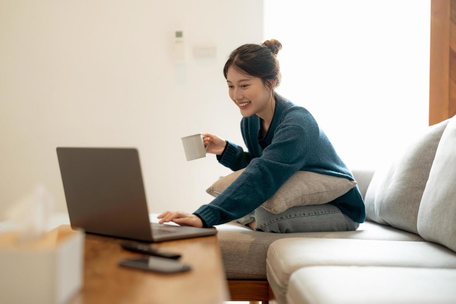 una joven asiática tranquila que trabaja en una laptop sentada en un sofá acogedor, una chica feliz navegando por Internet o comprando en línea durante el fin de semana soleado en casa, relajándose en el sofá con la computadora foto