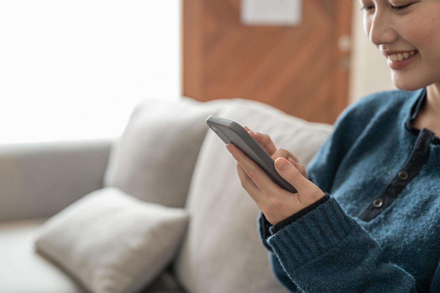 Cierra a una joven asiática alegre usando un teléfono móvil mientras te sientas en un sofá en casa foto